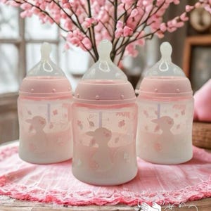 Op de afbeelding: Drie roze babyflessen met doorzichtige spenen en een lammetjesontwerp. De flessen staan op een roze kanten doek, met roze bloemen op de achtergrond. De tekst "Stacey's Reborn Nursery" is zichtbaar.