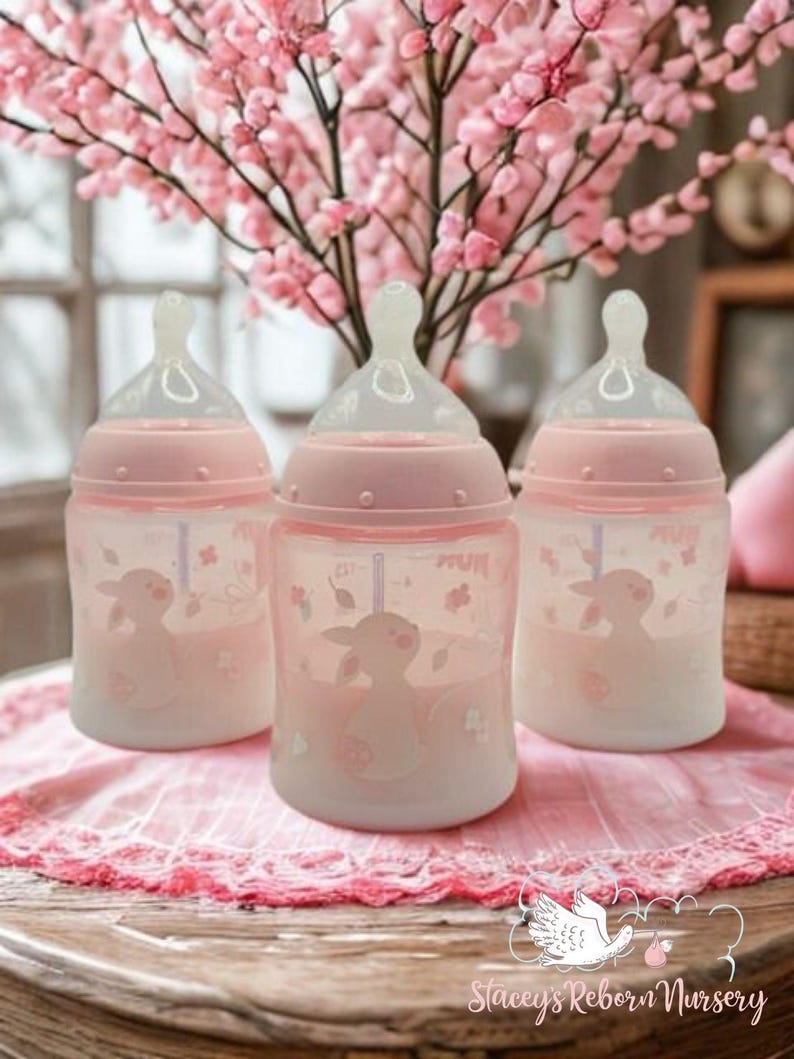 Op de afbeelding: Drie roze babyflessen met doorzichtige spenen en een lammetjesontwerp. De flessen staan op een roze kanten doek, met roze bloemen op de achtergrond. De tekst "Stacey's Reborn Nursery" is zichtbaar.