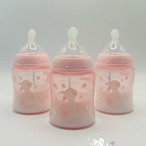 Op de afbeelding: Drie roze babyflessen met doorzichtige spenen. Elke fles heeft een roze lammetjesillustratie en bloemaccenten. De flessen hebben maatstreepjes en zijn ontworpen voor het voeden van baby's.