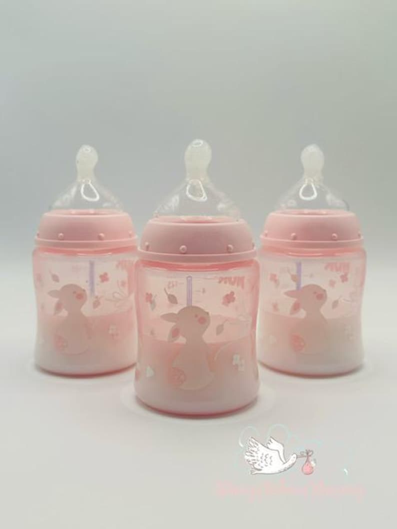 Op de afbeelding: Drie roze babyflessen met doorzichtige spenen. Elke fles heeft een roze lammetjesillustratie en bloemaccenten. De flessen hebben maatstreepjes en zijn ontworpen voor het voeden van baby's.
