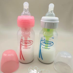 May include: Two clear baby bottles with pink and white caps, filled with a white liquid. One bottle has a pink cap and red accents, the other has a white cap and blue accents. The bottles are labeled "Dr. Brown's".