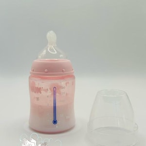Op de afbeelding: Een roze babyfles met een doorzichtige speen en een doorzichtige dop. De fles heeft een temperatuurindicator en meetmarkeringen. De fles is gevuld met een vloeistof. De merknaam "NUK" is zichtbaar op de fles.