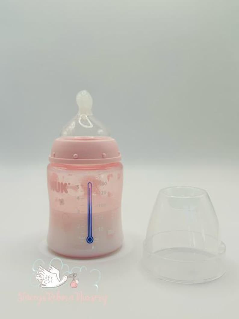 Op de afbeelding: Een roze babyfles met een doorzichtige speen en een doorzichtige dop. De fles heeft een temperatuurindicator en meetmarkeringen. De fles is gevuld met een vloeistof. De merknaam "NUK" is zichtbaar op de fles.