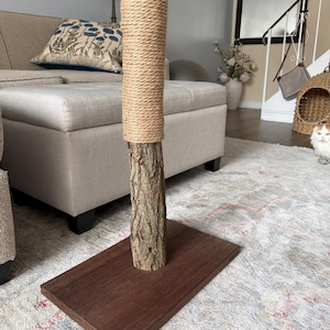 Peut inclure: Un griffoir pour chat avec une base en bois naturel et un poteau enroulé de corde de sisal. Le poteau est fait d'un tronc d'arbre et est conçu pour que les chats puissent gratter et grimper. La base est en bois foncé.