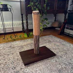 May include: A cat scratching post featuring a natural wood trunk wrapped in tan sisal rope. It is mounted on a dark brown rectangular base. The product is designed for cats to scratch and play.