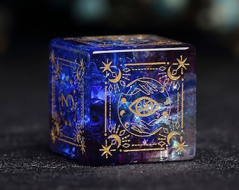 Puede incluir: Un dado de cristal azul y morado con detalles dorados. El dado presenta un diseño celestial con estrellas, lunas y un ojo.