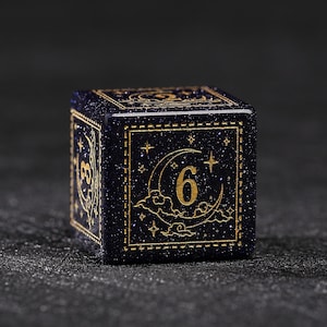 Könnte beinhalten: Ein dunkelblauer, glitzernder Würfel mit goldfarbenen Details. Der Würfel zeigt goldfarbene Zahlen und Himmelsmotive, darunter Monde und Sterne, in einem goldumrandeten Rahmen. Die Zahl 6 ist auf einer Seite zu sehen.