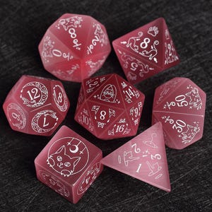 Könnte beinhalten: Ein Satz von sieben transluzenten roten Polyederwürfeln mit weißen geätzten Designs. Jeder Würfel zeigt Zahlen und Illustrationen zum Thema Katze. Die Würfel sind für Tabletop-Spiele konzipiert.
