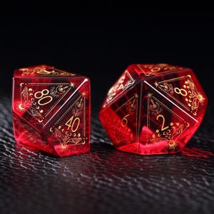 May include: Two translucent red polyhedral dice with gold ink engravings. The dice feature intricate celestial designs including hands, moons, and stars surrounding the numbers. One die shows the numbers 80 and 40, while the other shows 6 and 2.
