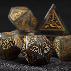 May include: Set of gemstone dice with gold-colored numbering and decorative designs. The dice are various polyhedral shapes, including a d20, d12, and d4. The dice have a dark, iridescent base color.