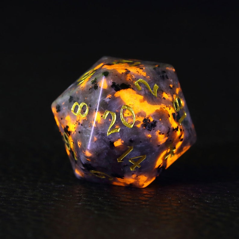 Peut inclure: Un d&eacute; d20 de couleur violette et noire mouchet&eacute; avec des chiffres dor&eacute;s. Le d&eacute; brille d'une couleur orange.