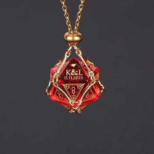 May include: A gold-colored chain necklace with a red, multi-sided pendant. The pendant has gold-colored accents and the text "K&L 11.11.2025" and the number 8. The necklace is displayed against a dark background.
