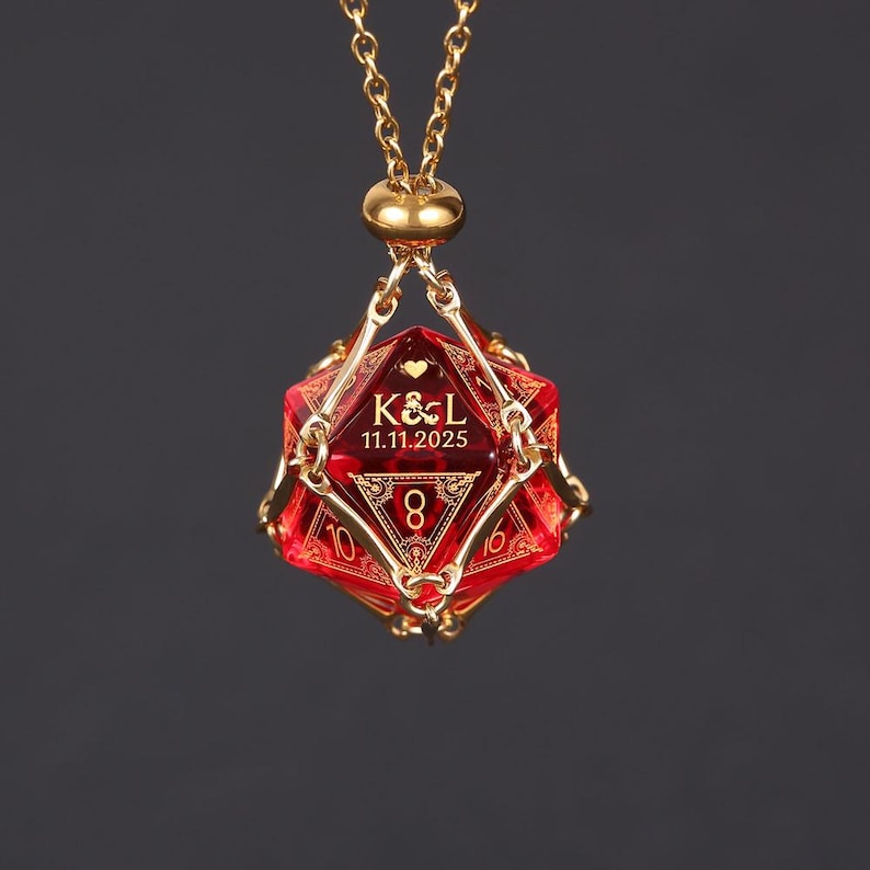 May include: A gold-colored chain necklace with a red, multi-sided pendant. The pendant has gold-colored accents and the text "K&L 11.11.2025" and the number 8. The necklace is displayed against a dark background.