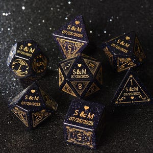 May include: A set of seven navy blue dice with gold-colored details. Each die features the initials "S & M" and the date "07/25/2025". The dice are in various geometric shapes, including a cube, a d20, and a tetrahedron.