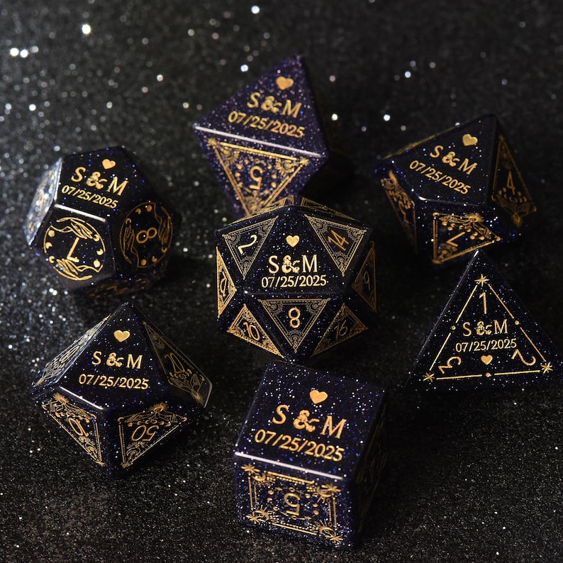 May include: A set of seven navy blue dice with gold-colored details. Each die features the initials "S & M" and the date "07/25/2025". The dice are in various geometric shapes, including a cube, a d20, and a tetrahedron.