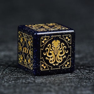 May include: A six-sided die in deep blue with a glittery finish, featuring gold-colored engravings on each face. One side displays a stylized octopus-like creature, while another side shows a geometric pattern with a central letter m symbol.