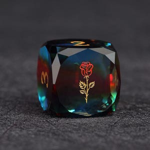 Op de afbeelding: Een facetgeslepen, iriserende dobbelsteen met een rozenafbeelding en gouden cijfers. De dobbelsteen is voornamelijk blauwgroen en rood, met gouden cijfers en een gouden rozenontwerp. De dobbelsteen ligt op een donkere ondergrond.