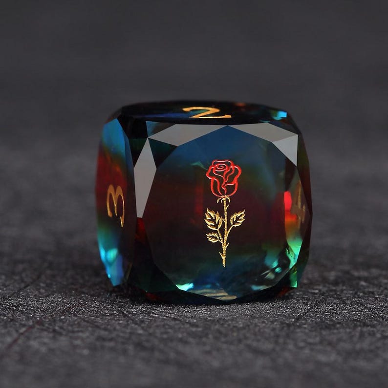 Op de afbeelding: Een facetgeslepen, iriserende dobbelsteen met een rozenafbeelding en gouden cijfers. De dobbelsteen is voornamelijk blauwgroen en rood, met gouden cijfers en een gouden rozenontwerp. De dobbelsteen ligt op een donkere ondergrond.