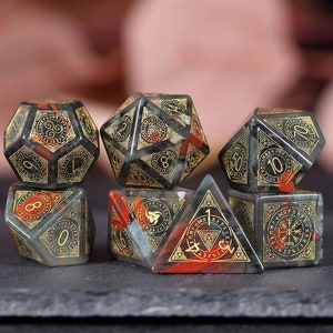 May include: Set of seven black, red, and gold polyhedral dice with intricate designs and runes. The dice are made of natural stone and have a speckled appearance.