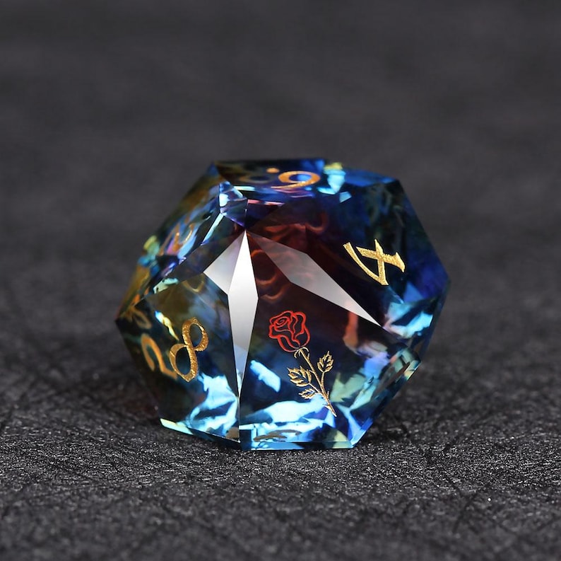Op de afbeelding: Een veelzijdige, blauwe en gouden D20 dobbelsteen met een roze afbeelding. De dobbelsteen heeft gouden cijfers en een heldere, edelsteenachtige uitstraling. De roos is rood met groene bladeren.