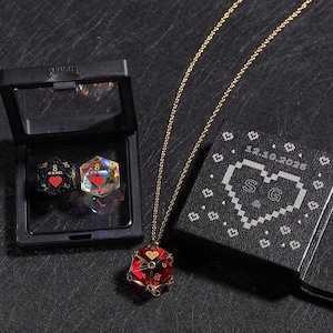 May include: A gold chain necklace with a red, gold, and clear glass dice pendant. The dice has a heart on one side and the initials "SG" on another. The necklace is displayed with a black box containing two dice, one black and one clear.