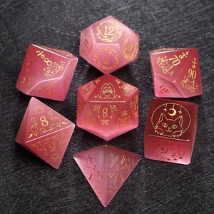 May include: Set of seven pink and gold dice with cat illustrations. The dice are in various shapes, including a d4, d6, d8, d10, d12, and d20.