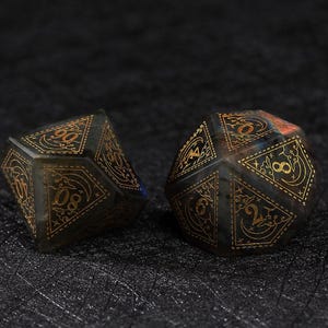 May include: Two polyhedral dice with gold-colored numbers and decorative designs. The dice are dark gray with a hint of blue and orange. The dice are for tabletop games.