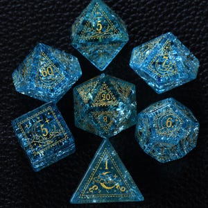 Puede incluir: Un juego de siete dados poliédricos de color azul translúcido con purpurina interna y tinta dorada. Cada dado presenta intrincados grabados celestiales de lunas, nubes y estrellas alrededor de los números, incluyendo un d20, d12, d10, d8, d6 y d4.