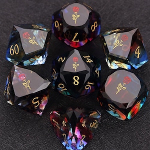 Op de afbeelding: Set van zeven polyhedrale dobbelstenen met een donkere, iriserende afwerking. Elke dobbelsteen heeft gouden cijfers en een rood rozenembleem. De dobbelstenen zijn ontworpen voor tabletop-rollenspellen en hebben verschillende geometrische vormen.