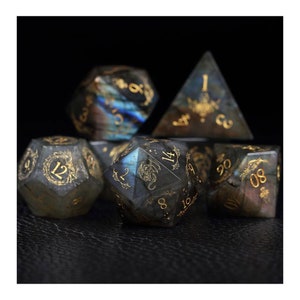 Könnte beinhalten: Satz von sieben schwarzen und goldenen Edelsteindice mit einem Drachenmotiv. Die Würfel sind aus Labradorit und haben eine goldene Metallic-Oberfläche.