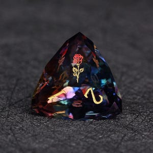 Op de afbeelding: Een veelkleurige, facetgeslepen polyhedrale dobbelsteen met een gouden roos en het nummer 2. De dobbelsteen heeft een iriserend uiterlijk met hints van rood, blauw en paars. Geschikt voor tabletop games.