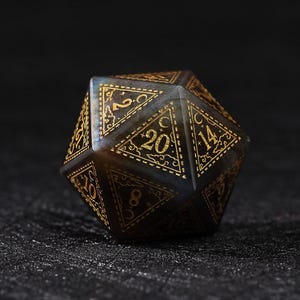 May include: A multi-sided, dark-colored die with gold-colored numbers and decorative accents. The die is a twenty-sided shape, with each side displaying a number from 1 to 20. The die is set against a dark background.