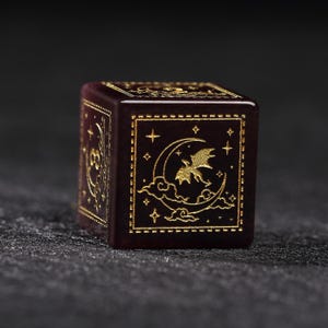May include: A burgundy-colored, six-sided die with gold-colored etched designs. Each face features a different celestial design, including moons, stars, and clouds. The die is a cube shape and appears to be made of a solid material.