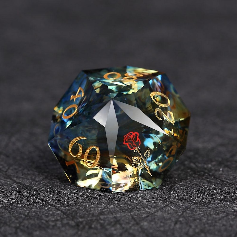 Op de afbeelding: Een veelzijdige, iriserende D60 dobbelsteen met gouden cijfers en een rode roosafbeelding. De dobbelsteen heeft een blauw-gouden kleurenschema, met een helder, geometrisch ontwerp. De getallen zijn 20, 40 en 60.