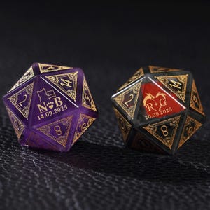 May include: Two twenty-sided dice with gold filigree numbering and borders. The purple die features a cat silhouette, the letters N B, and the date 14.09.2025. The black die features a dragon silhouette, the letters R + G, and the date 20.09.2025.