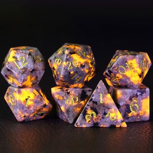 Peut inclure: Ensemble de sept d&eacute;s poly&eacute;driques en pierre translucide, mouchet&eacute;e, grise et noire avec des chiffres dor&eacute;s. Les d&eacute;s brillent de l'int&eacute;rieur d'une lumi&egrave;re orange chaude.