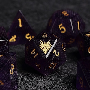 Könnte beinhalten: Ein Satz violetter und goldener polyedrischer Würfel für Tischspiele. Die Würfel haben ein strukturiertes, schuppenartiges Muster und goldene Zahlen. Ein Würfel zeigt ein goldenes Drachenemblem. Das Set enthält verschiedene Formen, wie z. B. einen d20, d12 und d4.