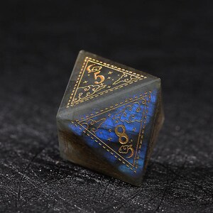 May include: A multi-sided, gemstone die with gold-colored numerals and decorative patterns. The die has a dark, iridescent base with blue and gold accents. The numbers 5 and 8 are visible on the faces of the die.