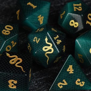 May include: A set of polyhedral dice in a deep emerald green with a subtle snakeskin texture pattern. Each die features gold-coloured numbers and a gold snake graphic on the highest face. The dice have a smooth, polished finish and sharp edges.