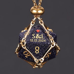 May include: A gold-colored necklace featuring a blue-speckled, multi-sided die pendant. The die displays the initials "S & L", a heart, and the date "12.12.2024" in gold lettering. The number "8" is also visible.