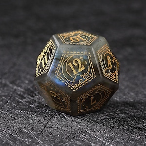 May include: A multi-sided, gemstone die with gold-colored numerals and decorative accents. The die is a mix of gray and blue with a polished finish. The numbers are in a classic font, and the die is likely used for tabletop gaming.
