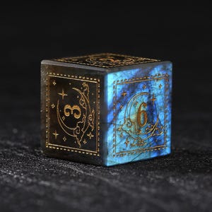 May include: A square, gemstone die with gold-colored numerals and celestial designs. The die features a blue and black color scheme with a moon and star motif. The number "6" is visible on one face.