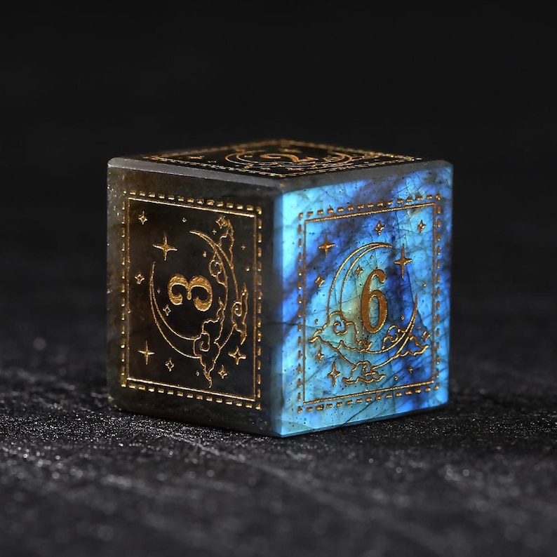 May include: A square, gemstone die with gold-colored numerals and celestial designs. The die features a blue and black color scheme with a moon and star motif. The number "6" is visible on one face.