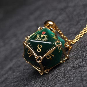 May include: A gold necklace with a green, eight-sided dice pendant. The dice has gold accents and the inscription "K & E 02.09.2025" on one side.
