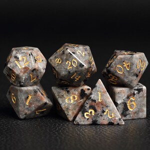 Peut inclure: Ensemble de sept d&eacute;s mouchet&eacute;s noirs et gris avec des chiffres dor&eacute;s. Les d&eacute;s sont de formes vari&eacute;es, notamment un cube, une pyramide et un dod&eacute;ca&egrave;dre.