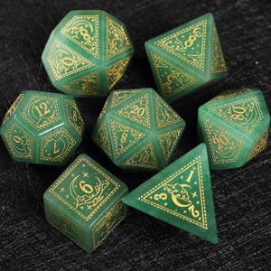 Puede incluir: Conjunto de siete dados poliédricos verdes con números y diseños decorativos dorados. Los dados, en formas como d4, d6, d8, d10, d12, d20 y un dado de porcentaje, se muestran sobre un fondo oscuro. Cada dado tiene detalles dorados.