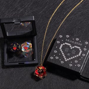 May include: A gold chain necklace with a red, faceted pendant shaped like a die. The pendant has gold accents and the initials "SG". A black box with a pixelated heart and the initials "S & G" is next to the necklace. Two dice are in a black case.