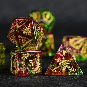 Pode incluir: Um conjunto de dados poliédricos feitos de resina translúcida com uma mistura de cores vermelha e verde. Cada dado apresenta gravuras douradas intrincadas de rosas e espadas, com números dourados claros indicando os valores em cada face.
