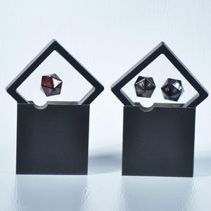 Op de afbeelding: Twee zwarte display cases met elk een polyhedrale dobbelsteen. E&eacute;n case heeft een rode en zwarte dobbelsteen, de andere heeft twee veelzijdige dobbelstenen met gouden cijfers. De cases hebben een zwarte vierkante basis en een zwart schuin frame.