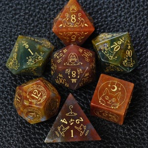 May include: A set of seven polyhedral dice made from mottled red, orange, and green stone. Each die features gold-colored engraved cat-themed illustrations, including paw prints, crescent moons, and feline faces, alongside standard numerical markings.